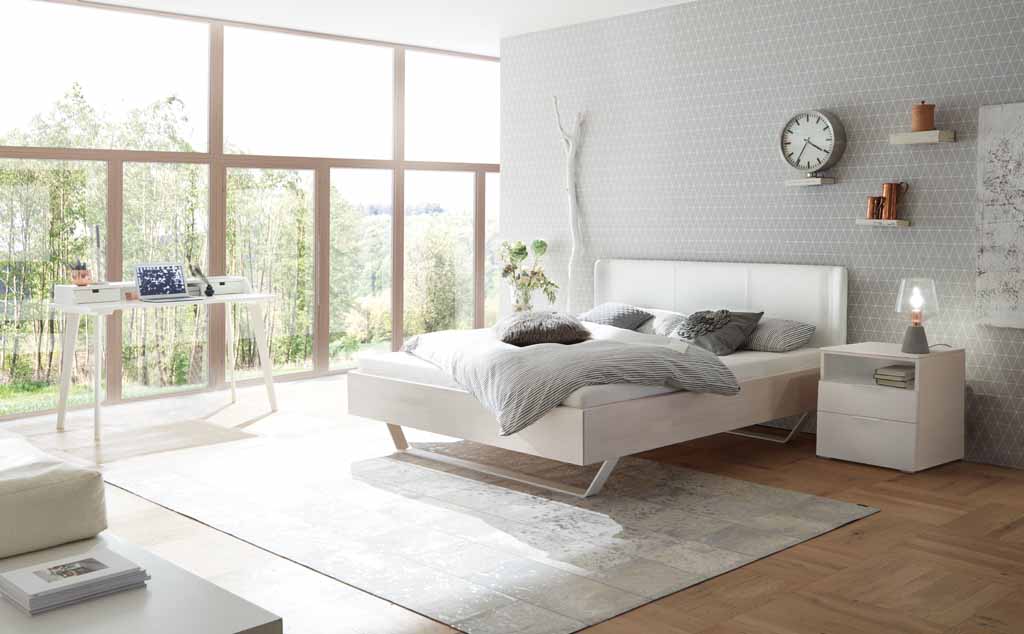 Hasena Wood-Line Bett | Buche weiß, lasiert | Kopfteil Stoff weiß Helles Schlafzimmer mit einem weißen Bett, Nachttisch, großem Fenster und dekorativen Details