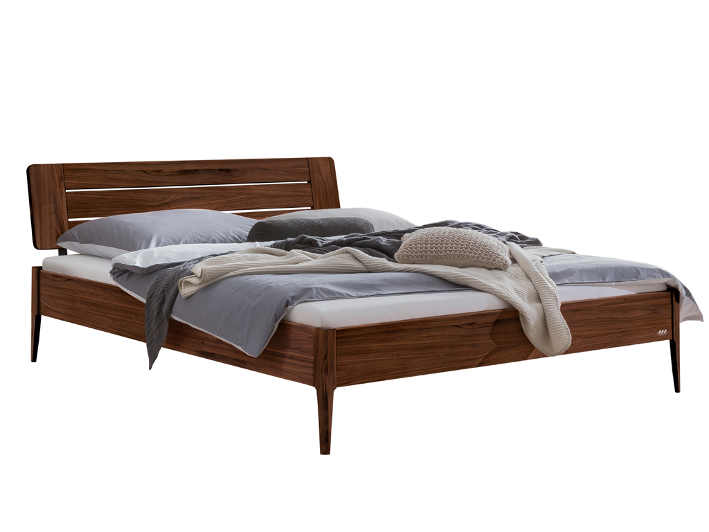 Eine Freisteller-Ansicht des Bettes "Busseto" mit einem Rahmen aus Nussbaumholz. Das Bett hat ein passendes Holzkopfteil mit horizontalen Lamellen und steht auf vier konischen Holzfüßen. Es ist mit grauer und grüner Bettwäsche dekoriert.