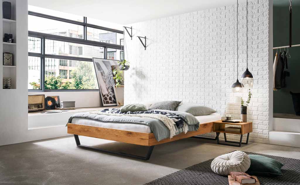 Hasena Trento - Bett Ostino | Wildeiche natur, geölt Helles Schlafzimmer mit einem modernen Holzbett, weißer Backsteinwand, großen Fenstern und gemütlichen Dekorationsgegenständen.