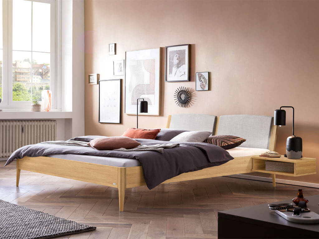 Ein stilvolles Schlafzimmer mit einem Hasena Pistoia Bett aus heller Eiche. Das Bett hat ein gepolstertes, hellgraues Kopfteil und einen passenden, schwebenden Nachttisch. Die Wände sind in einem hellen Lachston gestrichen.