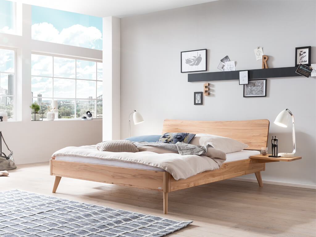 Ein Hasena Bett Caluso aus Wildbuche in einem hellen, modern eingerichteten Schlafzimmer mit großen Fenstern. Das Bett hat ein geschwungenes Kopfteil und schräg ausgestellte Beine.