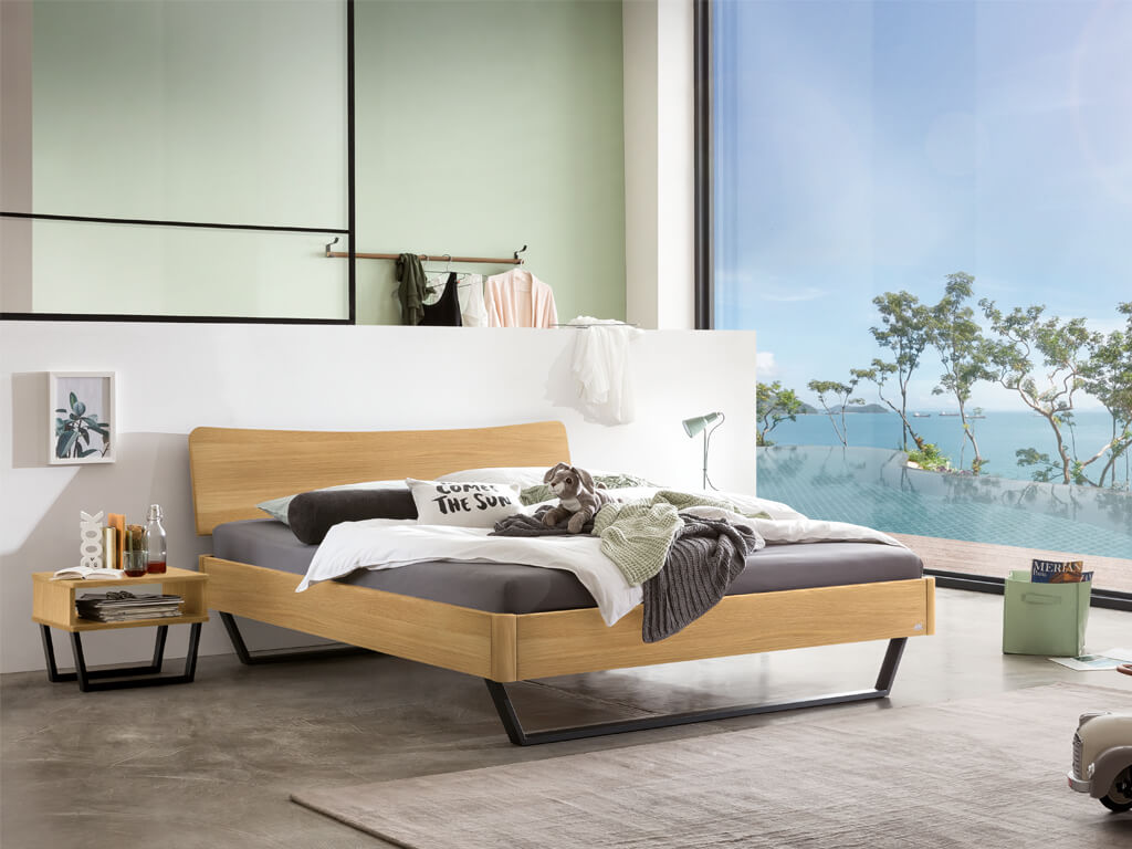 Ein Hasena Bettmodell Tresali aus heller Eiche, das in einem hellen, modern eingerichteten Schlafzimmer mit großen Fenstern steht.