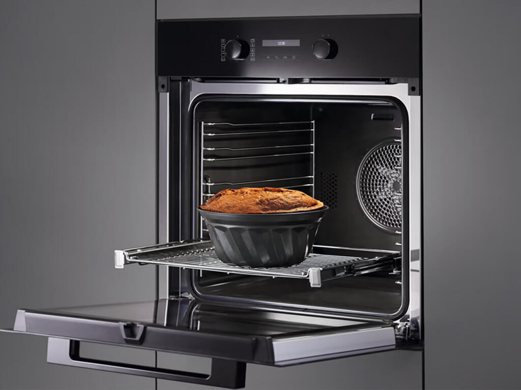 Eine Nahaufnahme des geöffneten Innenraums eines Miele-Backofens. Ein dunkler, runder Kuchen in einer Gugelhupf-Form steht auf einem ausziehbaren Rost in der Mitte des Backofens.