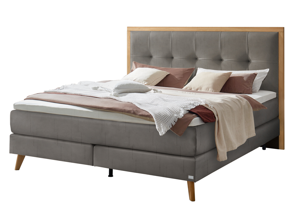 Ein Boxspringbett in einem samtigen Grauton mit einem Holzrahmen um das gepolsterte Kopfteil, freigestellt auf weißem Hintergrund.