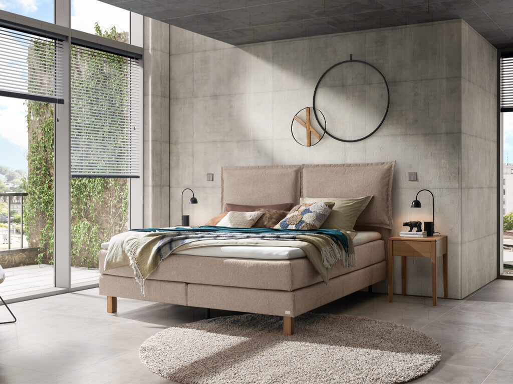 Ein modernes Schlafzimmer mit einem sandfarbenen Hasena Boxspringbett "Pronto Bonita" und einem Beton-Look an den Wänden, aufgenommen von einem anderen Blickwinkel.