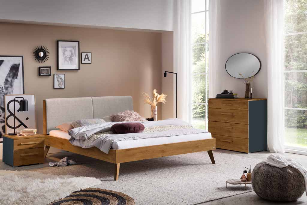Gemütliches Schlafzimmer mit Holzbett, Nachttischen, Kommode und dekorativen Elementen vor großen Fenstern
