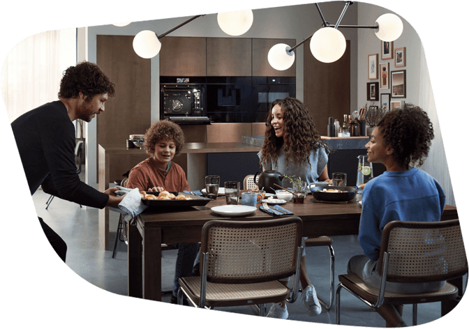 Eine Familie mit zwei Kindern sitzt fröhlich an einem Esstisch in einer modernen Küche. Ein Mann serviert das Essen, während alle lächeln.