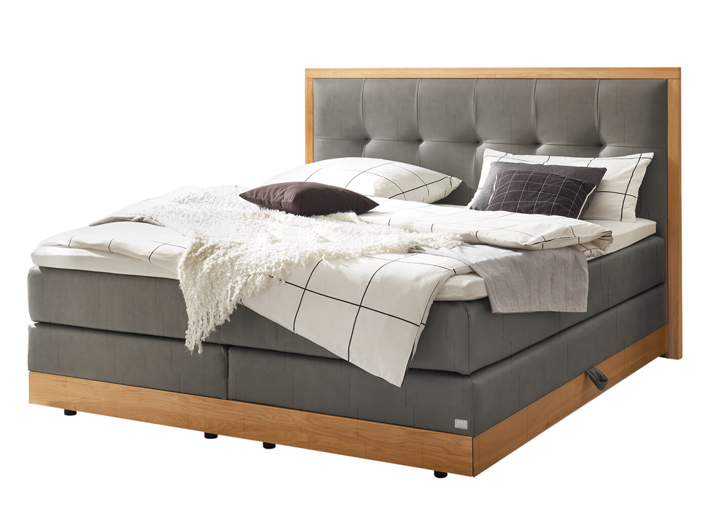 Modernes Boxspringbett "Hasena Pronto Alba" mit hellem Holzrahmen, grauem Stoffbezug und getuftetem Kopfteil. Weiße, graue und schwarze Bettwäsche, Kissen und eine weiße Decke.