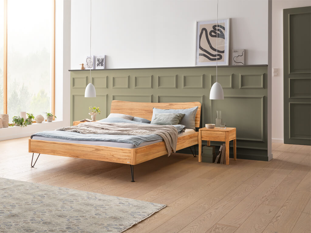 Hasena -  Lounge Bett Locarno | Holzkopfteil Wave | Metallfuß Locito | versch. Größen & Farben konfigurierbar Ein Hasena Bett Locarno aus Wildbuche, in einem hellen Schlafzimmer mit minimalistischer Dekoration und Haarnadel-Beinen in Anthrazit. Auf der rechten Seite steht ein passender Nachttisch.