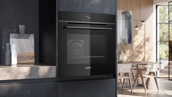 Eleganter, eingebauter, schwarzer studioLine Backofen in einer modernen, dunklen Küchenzeile mit einem Essbereich im Hintergrund.
