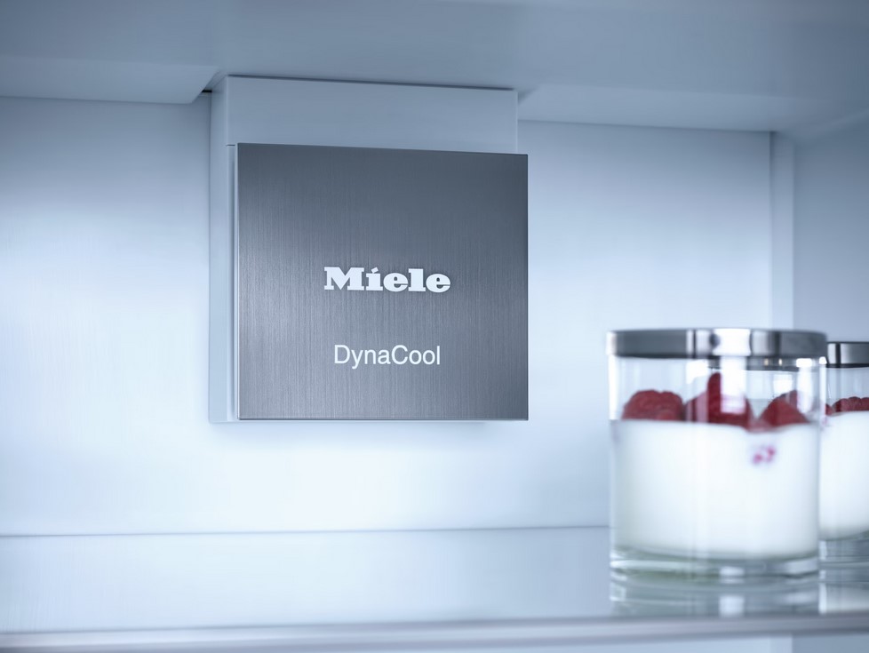 Detailansicht des Innenraums eines Miele Kühlschranks, die das silberne "Miele DynaCool"-Schild an der Rückwand zeigt. Auf einem Glasregal im Vordergrund stehen zwei Gläser mit Joghurt und Himbeeren.