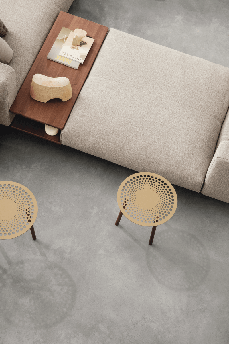 Ein Wohnzimmer mit einem beigen Sofa, einer hölzernen Tischablage mit Büchern und Dekoration und zwei runden Metallhockern auf einem grauen Boden.