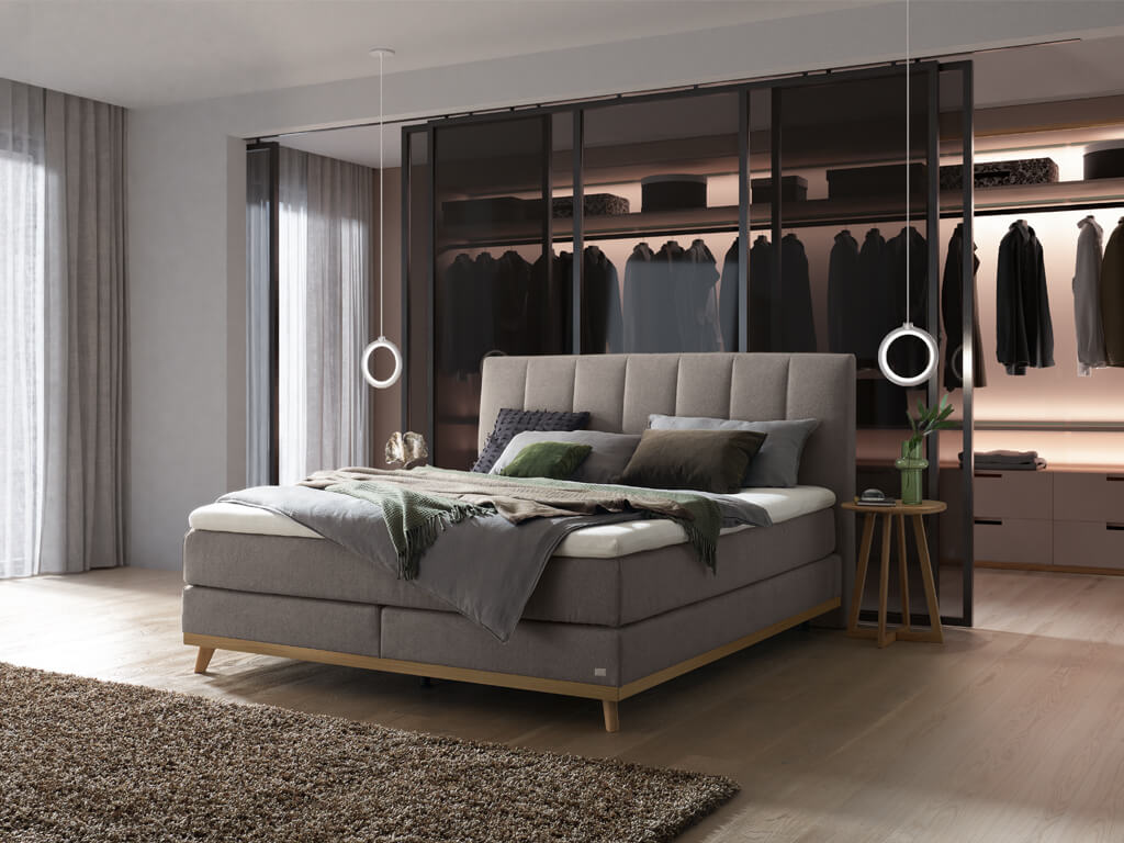 Ein Hasena Boxspringbett Pronto Stefania Roy mit grauem Stoffbezug in einem Schlafzimmer mit einem modernen, offenen Kleiderschrank.