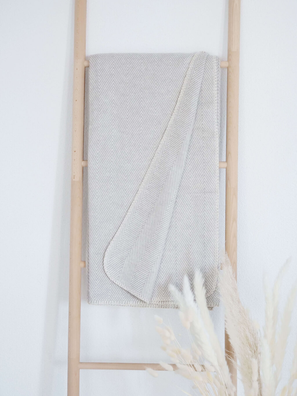 RIEMA. FINN – Wohndecke, Bio-Baumwolle - Fischgrät | 140 x 200 cm | hellgrau Eine beige Decke mit Fischgrätmuster hängt auf einer Holzleiter, im Vordergrund dekorative Gräser