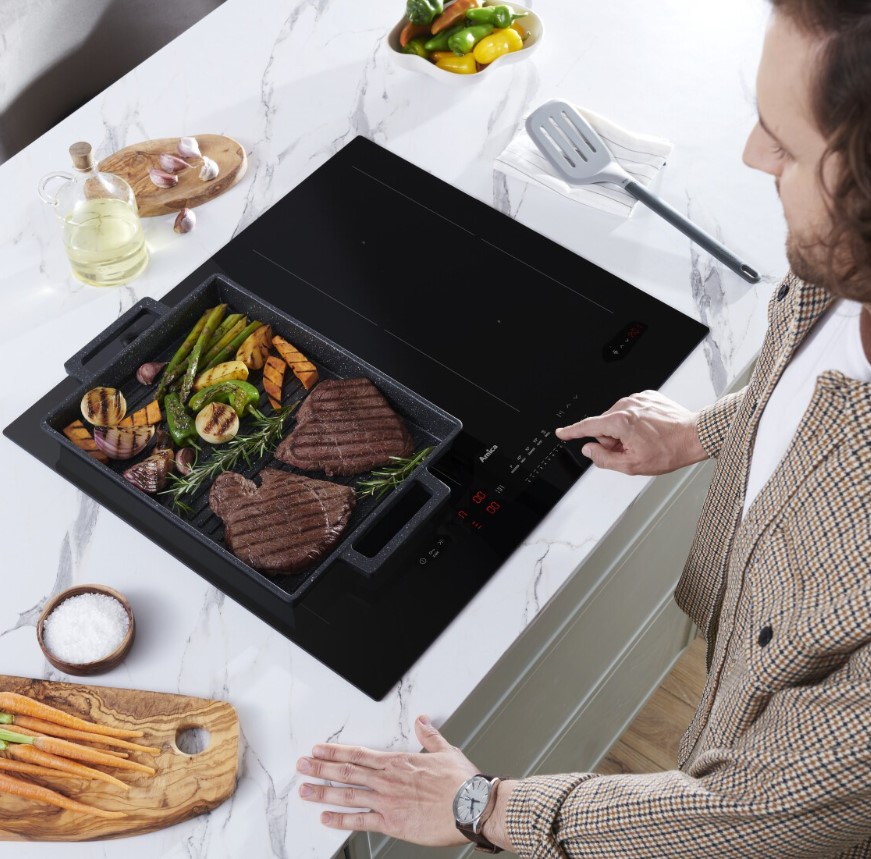 Ein Mann steht über einem modernen, schwarzen Amica Induktionskochfeld und bedient die Touch-Steuerung, während in einer Grillplatte auf dem Kochfeld Fleisch und Gemüse braten.