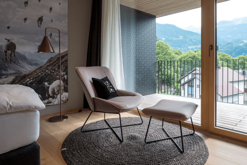 Innenraum eines modernen Wohnzimmers mit Sessel, Fußhocker, Stehlampe und Balkonblick auf die Berge