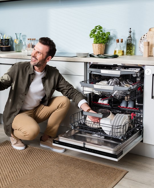 Ein lächelnder Mann in einer beigen Hose und einem olivgrünen Hemd hockt vor einer geöffneten Amica Spülmaschine und räumt Geschirr ein.