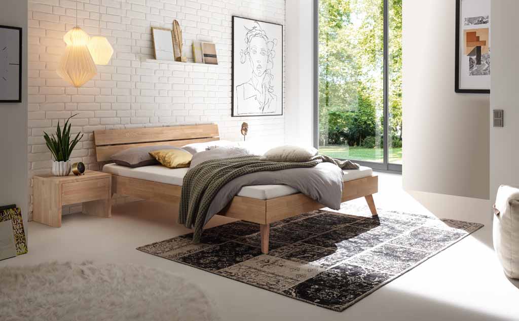 Hasena Trento - Bett Ortona | Eiche bianco, geölt Gemütliches Schlafzimmer mit einem natürlichen Holzbett, stilvollen Dekoelementen und einer großen Fensterfront mit Blick ins Grüne.