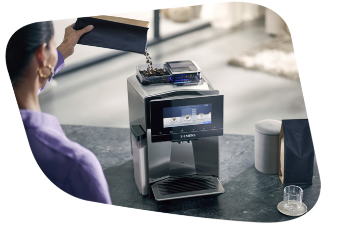 Eine Person füllt Kaffeebohnen aus einem schwarzen Beutel in das Bohnenfach eines Siemens Kaffeevollautomaten. Der Vollautomat hat ein Touch-Display und eine silberne Oberfläche.