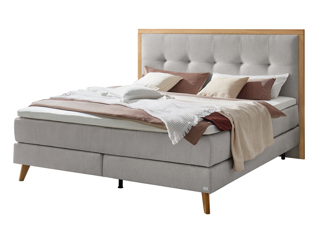 Ein Boxspringbett in hellem Grau mit einem Holzrahmen um das gepolsterte Kopfteil, freigestellt auf weißem Hintergrund.
