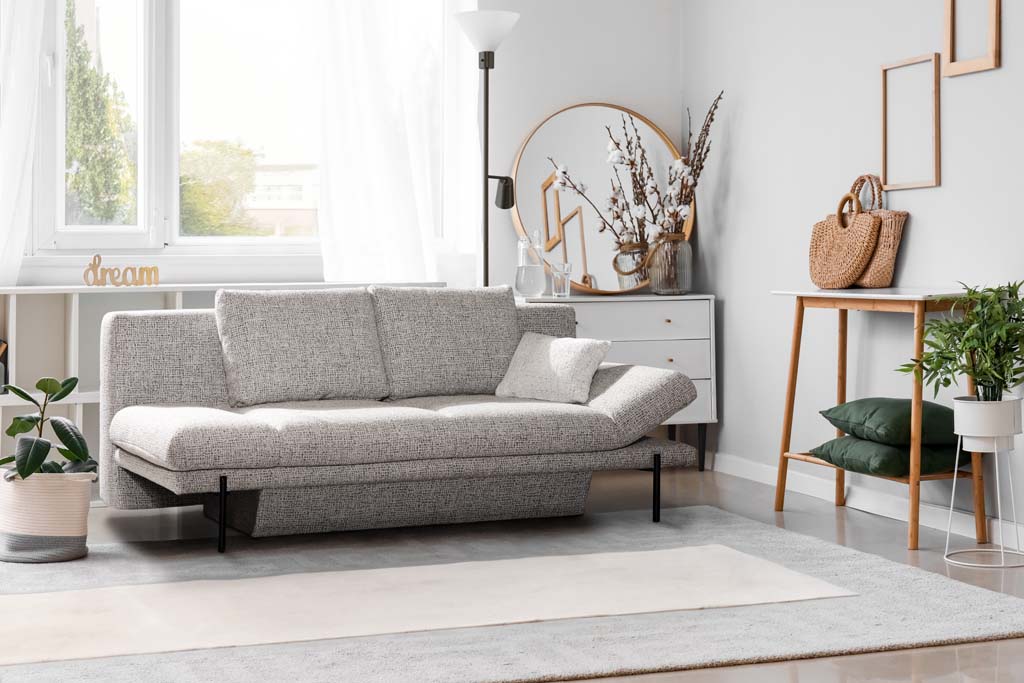 Restyl Vivien - Schlafsofa (Querschläfer) | grau | mit Bettkasten | 150 x 207 cm Wohnzimmer mit grauem Sofa, Pflanzen, Holzregal und Dekorationen in einem hellen, modernen Raum.