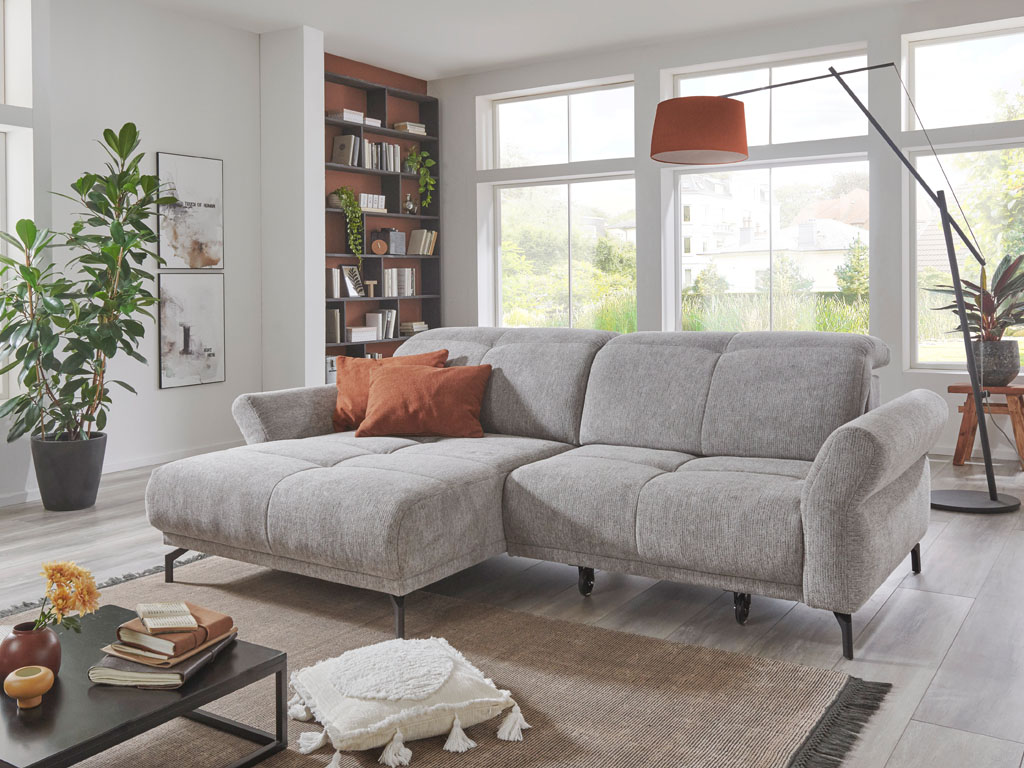 Poco Kingston Move - Funktionssofa Ein helles Wohnzimmer mit einem gemütlichen, grauen Ecksofa, dekorativen Kissen, einem modernen Teppich, einem Couchtisch mit Dekoartikeln und großen Fenstern.