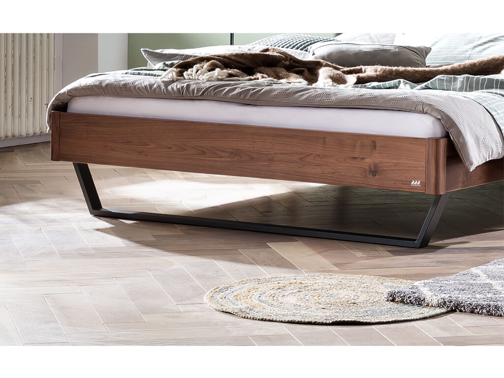 Nahaufnahme des Fußendes eines Bettes mit einem Rahmen aus dunkelbraunem Nussbaumholz. Das Bett steht auf einem schwarzen Metallgestell. Der Boden ist aus hellem Holz und es liegen zwei kleine Teppiche davor.