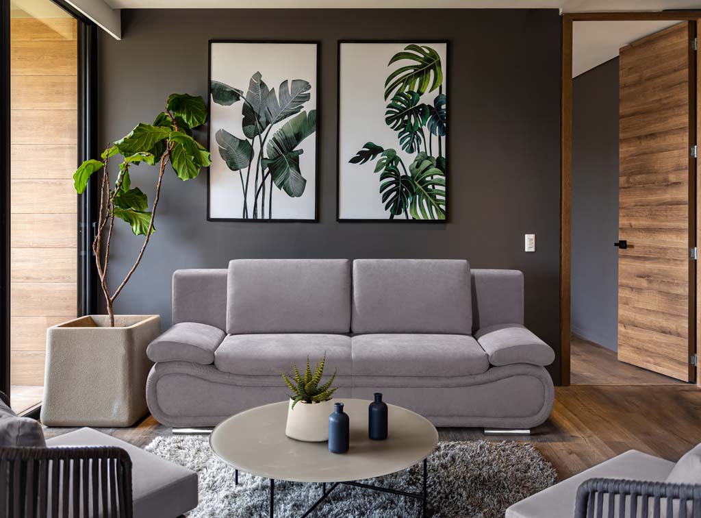 Ein modernes Wohnzimmer mit einem grauen Sofa, umgeben von grünen Pflanzen und stilvollen Wandbildern. Der Raum wirkt gemütlich und einladend.