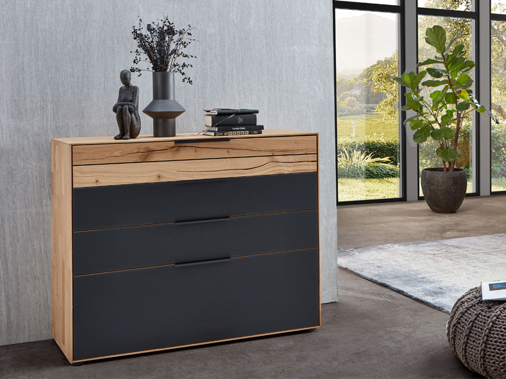 Wöstmann WSM 4300 - Kommode (2252), 3 Schubkästen, 1 Auszug | Wildeiche massiv | B: 121,9 cm | konfigurierbar Eine stilvolle Kommode aus Holz und Metall mit Dekorationsgegenständen darauf, in einem modernen Wohnzimmer mit großen Fenstern und Blick auf einen Garten.