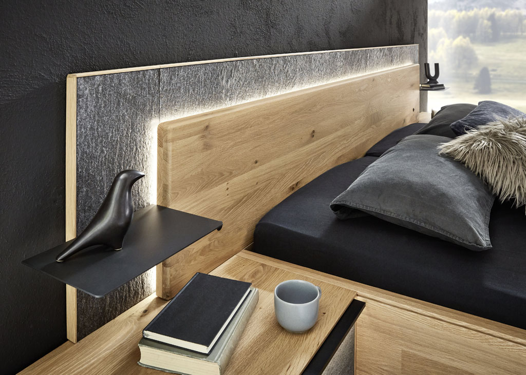 Detailaufnahme eines Bettes mit beleuchtetem Holzkopfteil und schwarzer Metallablage sowie einer grauer Kopfteilumrandung stilvoll dekoriert