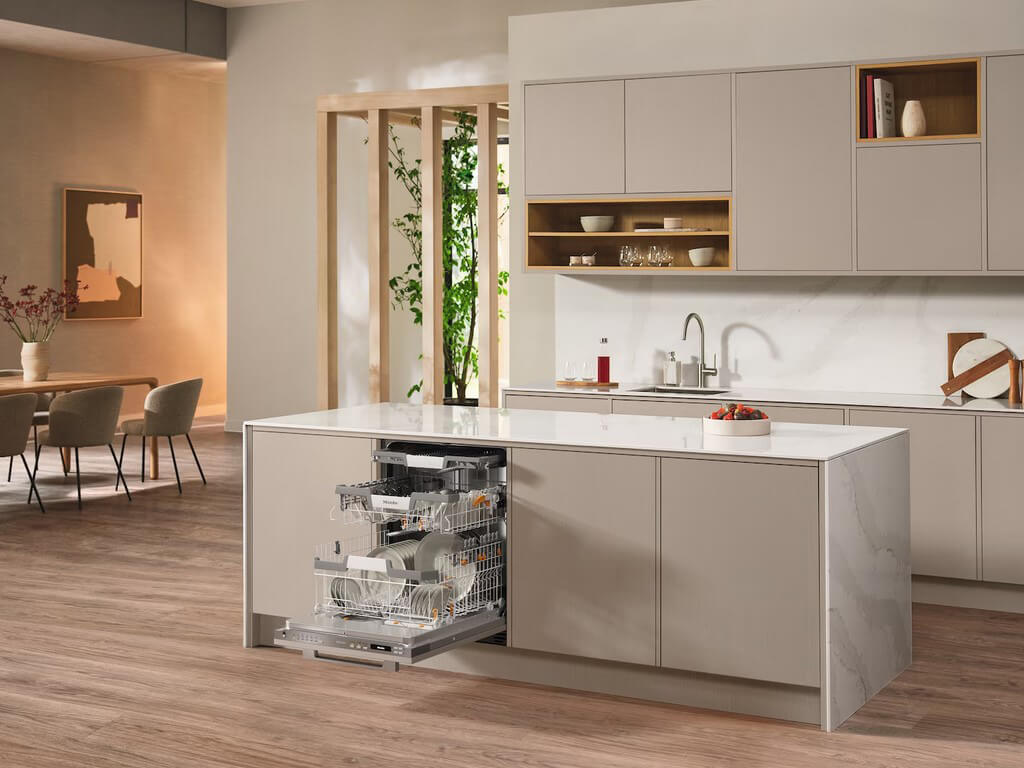 Eine moderne Küche mit einer geöffneten Miele Geschirrspülmaschine, die in die Kücheninsel integriert ist. Die Geschirrkörbe sind herausgezogen und mit Geschirr beladen.