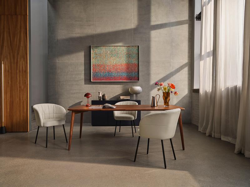 Ein moderner Essbereich mit einem langen Holztisch, umgeben von eleganten weißen Stühlen. An der Wand hängt ein buntes Kunstwerk, das dem Raum einen Farbakzent verleiht. Lichtdurchfluteter Raum mit großen Fenstern und geschmackvoller Dekoration.