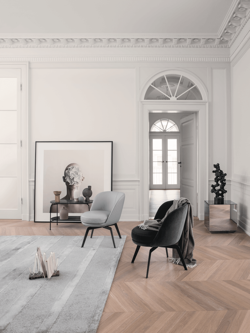 Ein stilvoll eingerichtetes Wohnzimmer mit zwei modernen Sesseln, einem Teppich und einem Kunstwerk, das an der Wand lehnt. Der Raum hat einen hölzernen Fischgrätboden und klassische Stuckverzierungen.