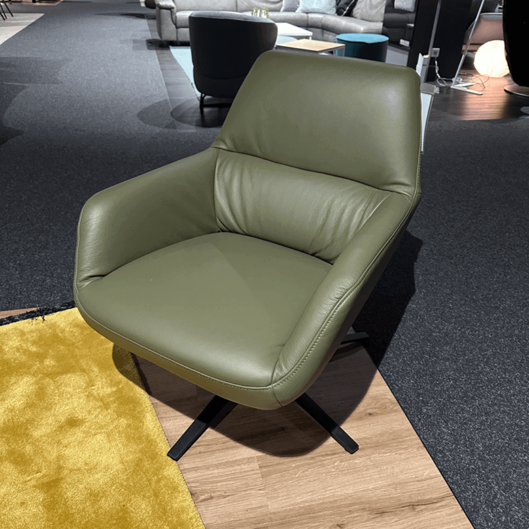 Himolla 7841 | Einzelsessel Ein moderner olivgrüner Ledersessel steht auf einem Holzboden. Der Stuhl hat einen schwarzen Metallfuß und befindet sich in einer wohnlichen Umgebung neben einem gelben Teppich.
