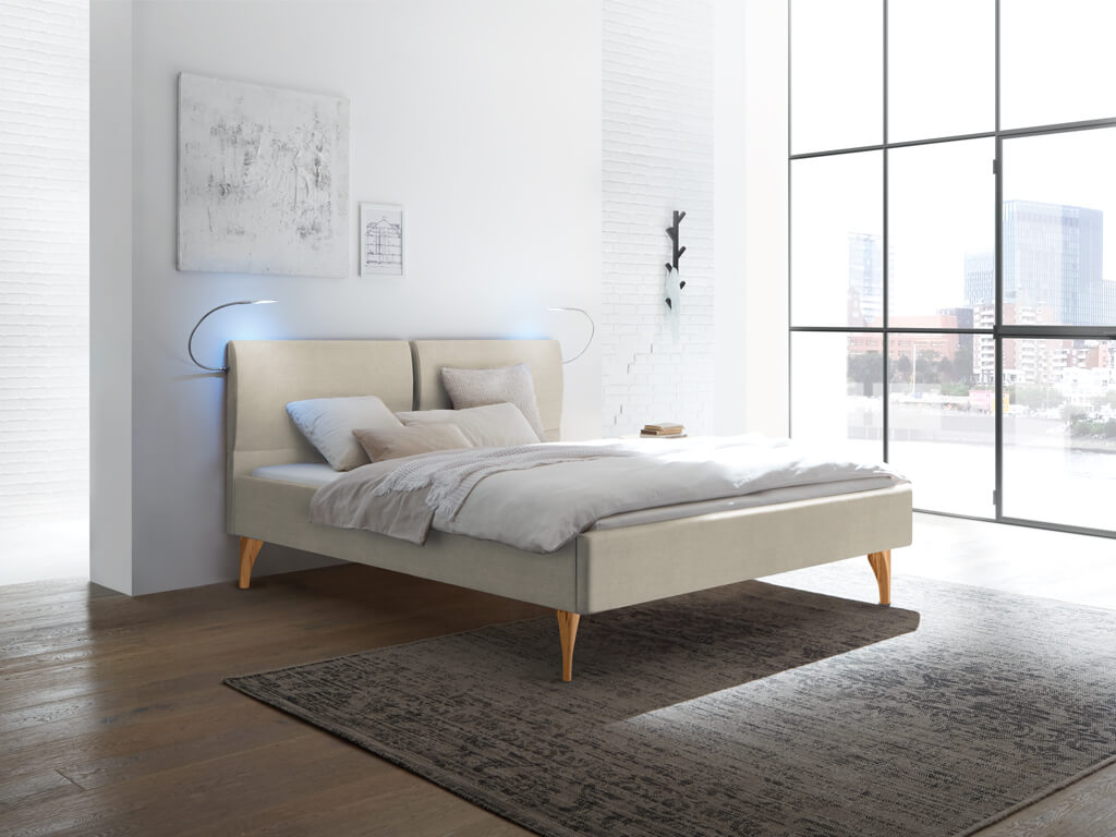 Ein beiges Polsterbett mit geteiltem Kopfteil und hellen Holzfüßen in einem minimalistischen Raum mit viel Tageslicht.