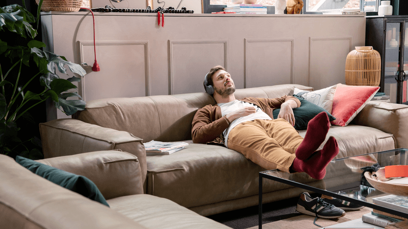 Ein Mann liegt entspannt auf einem beigen Ledersofa und hört Musik über Kopfhörer. Er trägt eine braune Strickjacke, ein weißes T-Shirt, beige Hose und rote Socken. Im Wohnzimmer sind Pflanzen und ein Glastisch zu sehen.
