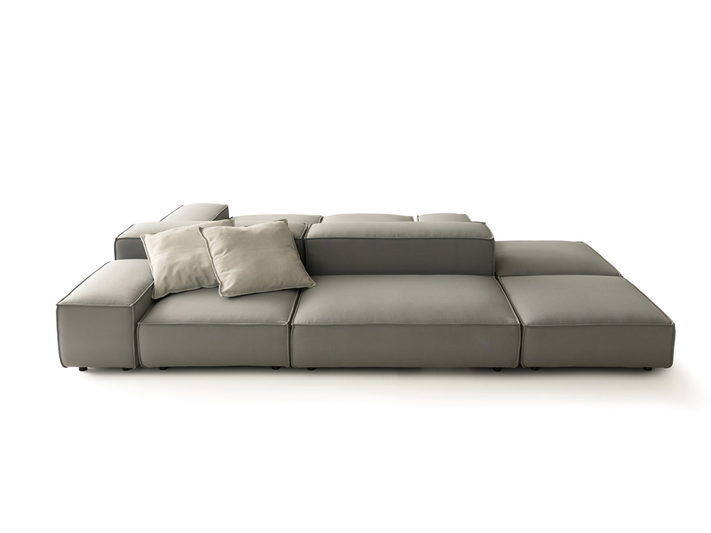 Ein modulares Outdoor-Sofa in Hellgrau, bestehend aus mehreren quadratischen Elementen, die zu einer länglichen Sitzlandschaft arrangiert sind. Zwei hellbeige Kissen liegen auf einem der Rückenelemente. Der Hintergrund ist weiß.