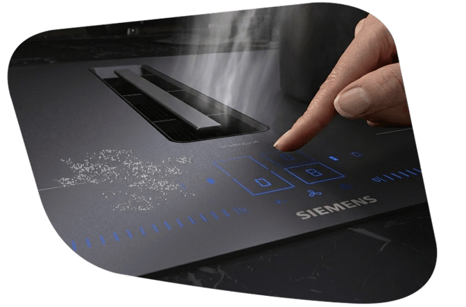 Nahaufnahme eines Siemens Induktionskochfelds mit matter Oberfläche und integriertem Dunstabzug. Ein Finger bedient das blau leuchtende Touch-Display, während Wasserdampf in den Abzug zieht.