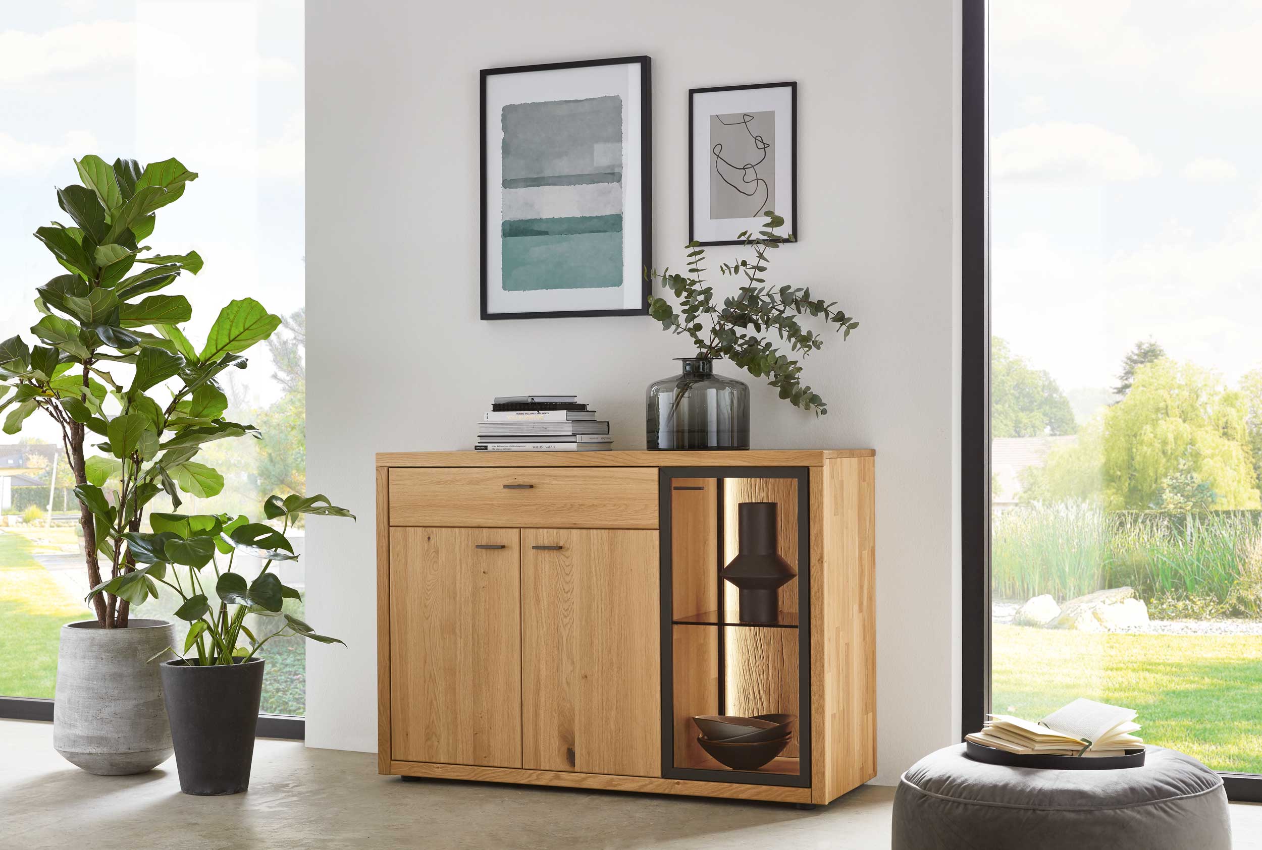 Wöstmann WM 2250 - Sideboard  |  Type 2253/2254 | Wildeiche massiv | B: 128,0 cm Helles Wohnzimmer mit großem Fenster, Echtholz-Kommode, zwei grünen Pflanzen, dekorativen Vasen und gerahmter Wandkunst