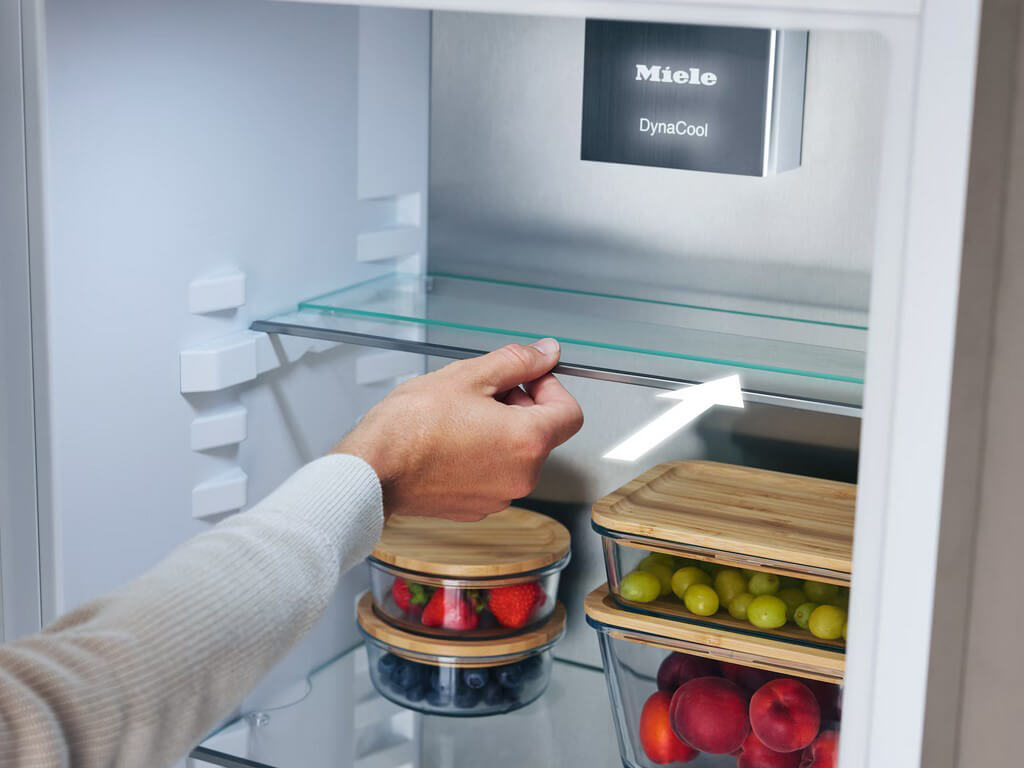 Eine Nahaufnahme des Innenraums eines Miele Kühlschranks. Eine Hand schiebt eine Glasablage nach hinten, um den Stauraum zu vergrößern.