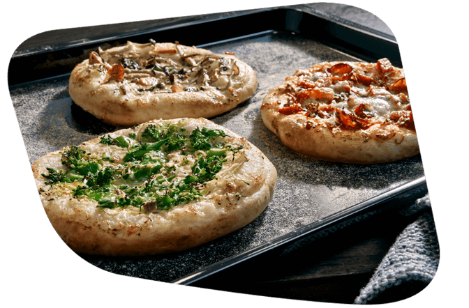 Ein Backblech mit drei kleinen Pizzen, belegt mit verschiedenen Zutaten wie Pilzen, Salami und Brokkoli.