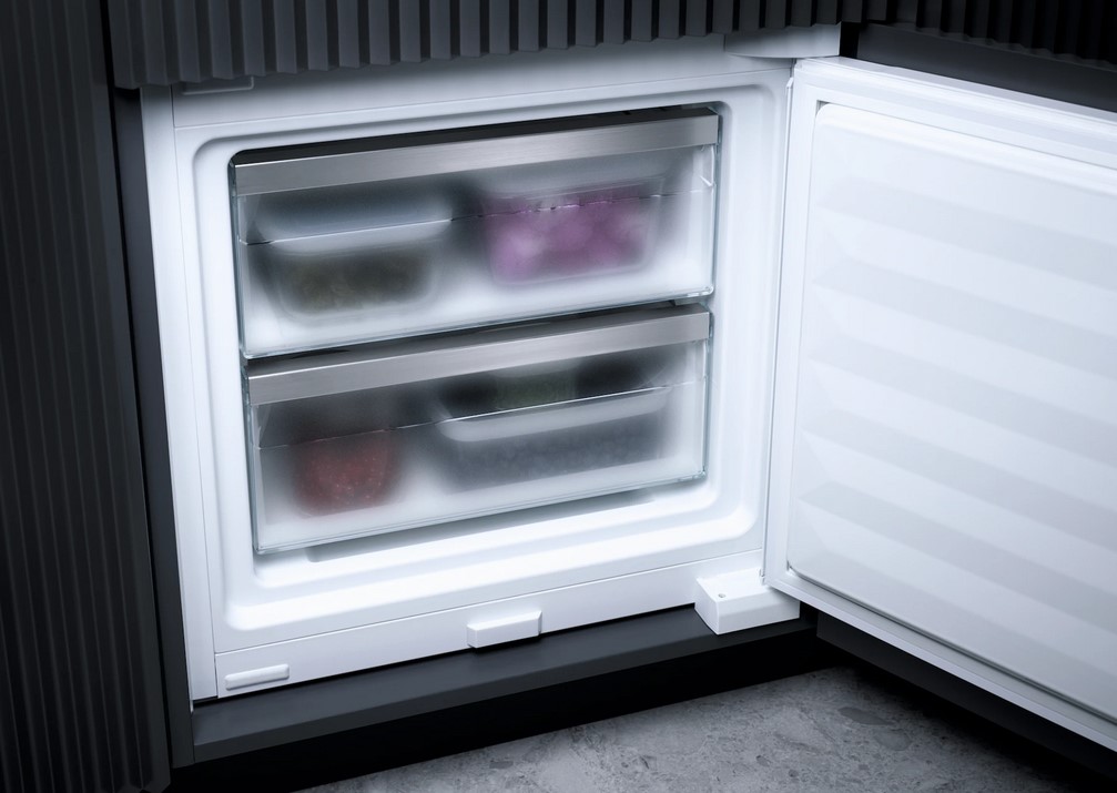  Nahaufnahme des geöffneten unteren Bereichs eines Miele Kühlschranks, der die zwei transparenten Gefrierschubladen zeigt, in denen gefrorene Lebensmittelbehälter zu erkennen sind. Der Kühlschrank ist in eine dunkle Küchenzeile integriert.