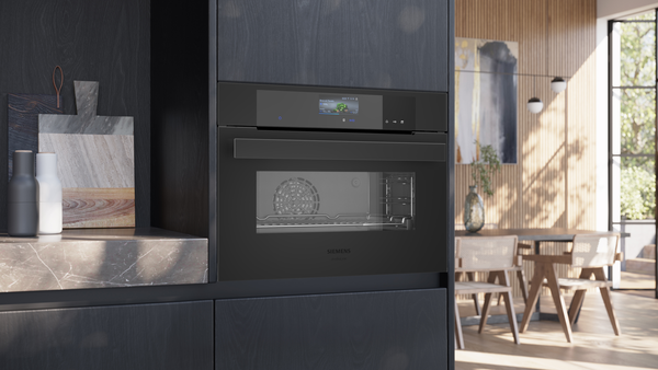 Ein kompakter, schwarzer Einbaubackofen in einer modernen, dunklen Küchenwand. Das aktive TFT-Display zeigt ein Programm für Brokkoli an; im Hintergrund ist ein heller Essbereich erkennbar.