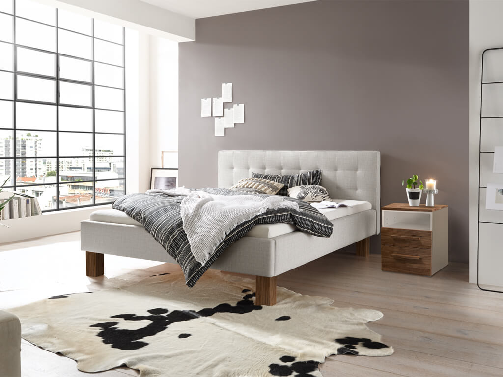 Ein hell eingerichtetes Schlafzimmer mit einem hellgrauen Hasena Noemi Bett mit gestepptem Kopfteil, dunklen Nussbaumholzbeinen und einem Kuhfell-Teppich.