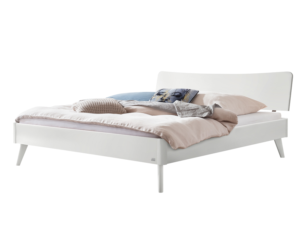 Ein freigestelltes, weiß lackiertes Hasena Bett Caluso. Das Bett hat ein geschwungenes Kopfteil, schräg ausgestellte Beine und ist mit Kissen und einer Decke dekoriert.