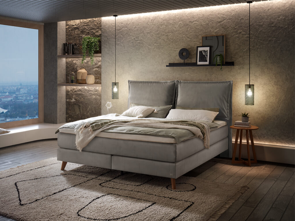 Ein Hasena Boxspringbett Pronto Ricarda Roy mit einem stahlgrauen Stoffbezug, das in einem Schlafzimmer mit einer strukturierten Wand steht.