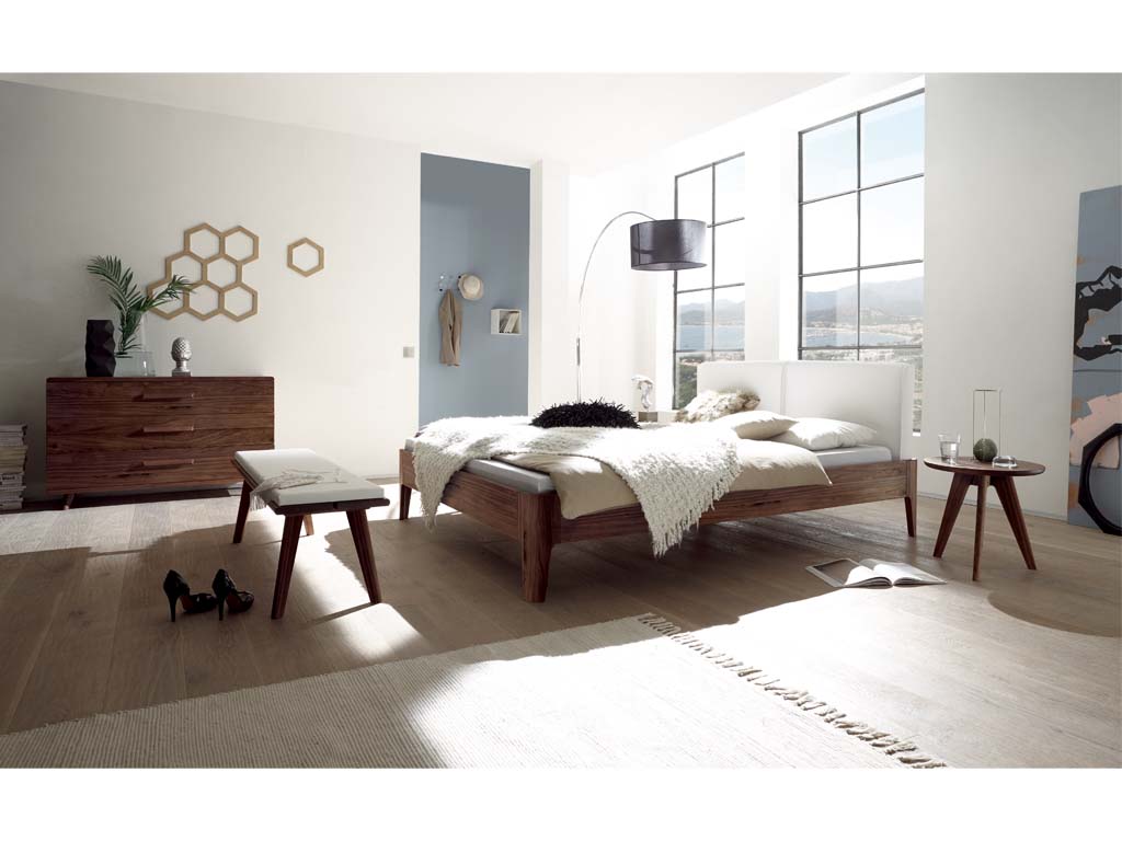 braunes Schlafzimmer in einem modernen Raum mit großen fenstern mit einem Bett einer Kommode mit drei Schubladen einem kleinen runden hocker einer holzbank mit weißem polster stilvoll dekoriert Hasena