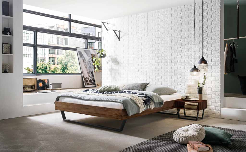 Hasena Trento - Bett Ostino | Nussbaum, geölt Ein stilvoll eingerichtetes Schlafzimmer mit einem Holzrahmenbett, weißer Ziegelwand und großen Fenstern, das Tageslicht hereinscheinen lässt. Bequeme Kissen und eine moderne Dekoration ergänzen den Raum.