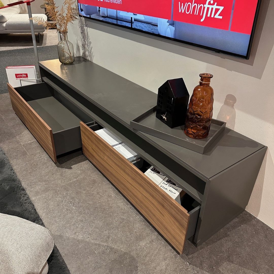 Moderner grauer TV-Schrank mit geöffneten Holzschubladen, dekoriert mit einer Vase, einem Tablett und einer kleinen schwarzen Skulptur. Im Hintergrund ist ein Bildschirm mit dem Logo von wohnfitz zu sehen.