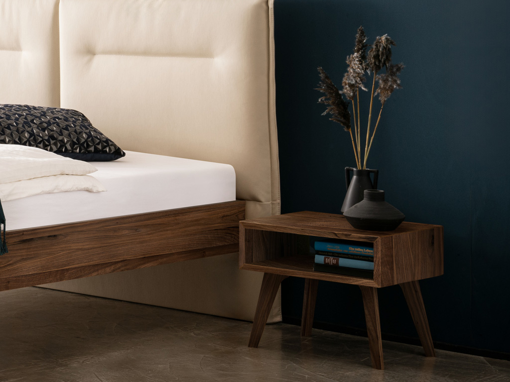 dunkelbrauner Nachttisch auf vier hohen beinen und in form eines offenen faches stilvoll dekoriert an einem bett stehend vor einer dunklen wand jaca von hasena